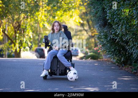 Das elfjährige Mädchen mit Ullrich kongenitaler Muskeldystrophie spielt in ihrem Rollstuhl in der Nähe ihres Heims Fußball Stockfoto