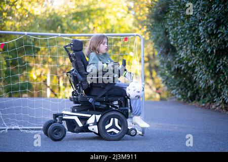 Das elfjährige Mädchen mit Ullrich kongenitaler Muskeldystrophie spielt in ihrem Rollstuhl in der Nähe ihres Heims Fußball Stockfoto