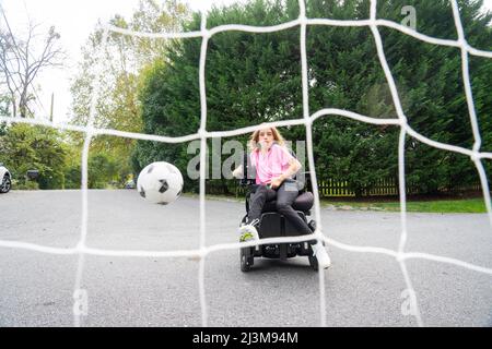 Das elfjährige Mädchen mit Ullrich kongenitaler Muskeldystrophie spielt in ihrem Rollstuhl in der Nähe ihres Heims Fußball Stockfoto