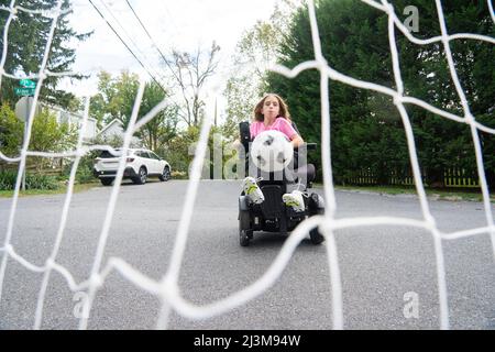 Das elfjährige Mädchen mit Ullrich kongenitaler Muskeldystrophie spielt in ihrem Rollstuhl in der Nähe ihres Heims Fußball Stockfoto