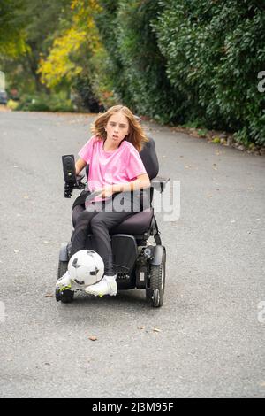 Das elfjährige Mädchen mit Ullrich kongenitaler Muskeldystrophie spielt in ihrem Rollstuhl in der Nähe ihres Heims Fußball Stockfoto