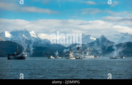 Zahlreiche Boote vor der zerklüfteten Küste von Alaska, mit schneebedeckten Berggipfeln im Hintergrund und Abgase, die von den Booten auf einem... Stockfoto