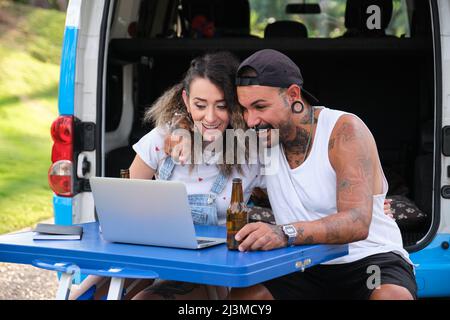 Das junge tätowierte Paar lächelt und sieht etwas im Laptop in der Nähe des Wagens. Stockfoto