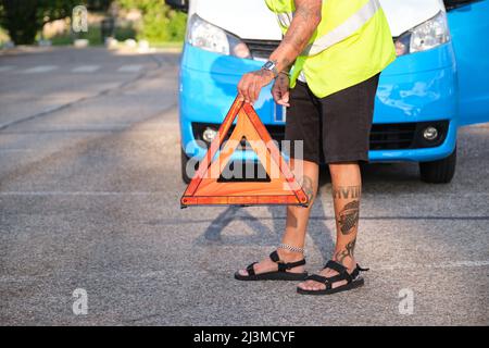 Nicht erkennbarer tätowierter Mann platziert ein reflektierendes Sicherheitsdreieck vor seinem Van. Stockfoto