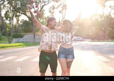Junge, coole tätowierte Paare brüllen, erheben ihre Arme und lachen mit einem Skateboard. Stockfoto