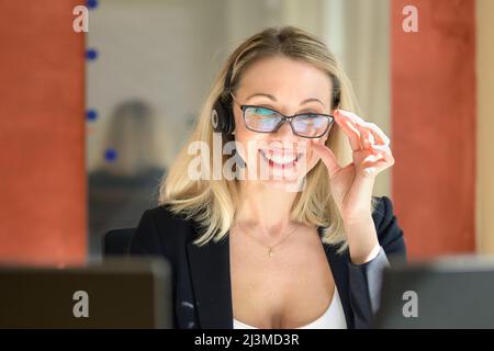 Lächelnde Geschäftsfrau in formeller Kleidung und einer Brille, die etwas liest, während sie im Büro mit dem Kunden über ein Headset spricht Stockfoto