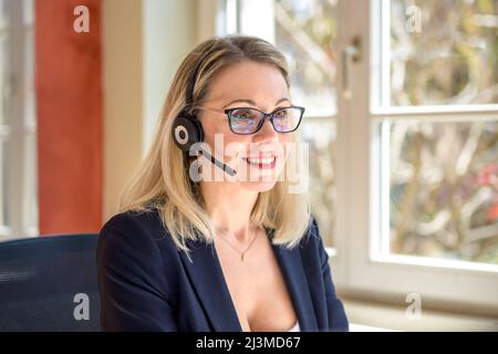 Lächelnde Geschäftsfrau, die formelle Kleidung und Brille trägt, während sie im Büro mit dem Kunden über ein Headset spricht Stockfoto