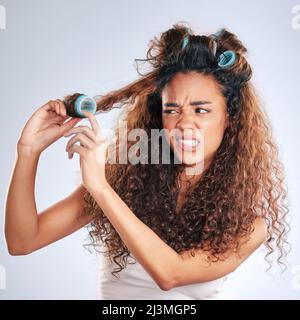 Das sollte funktionieren. Geschnittene Aufnahme einer attraktiven jungen Frau, die im Studio mit Lockenwicklern vor grauem Hintergrund kämpft. Stockfoto