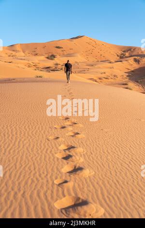 Ein Mann geht bei Sonnenuntergang eine orangefarbene Sanddüne hinauf, mit einer Spur sandiger Fußspuren im Vordergrund. Stockfoto