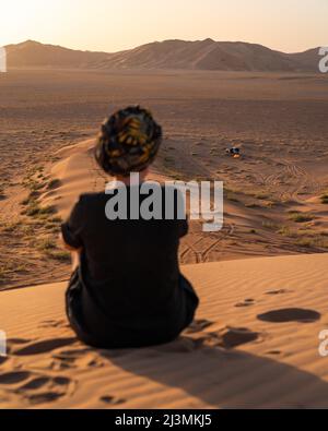 Eine Frau, die einen Turban trägt, sitzt bei Sonnenuntergang auf einer Sanddüne und blickt auf einen 4WD-Auto- und wilden Lagerplatz in der Wüste Rub al Khali (leeres Viertel) darunter. Stockfoto