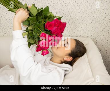 Ein junges und glückliches Mädchen liegt auf dem Bett und riecht mit geschlossenen Augen Rosen Stockfoto
