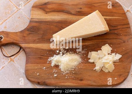 Nahaufnahme des ganzen Parmesankäses, der Holzspalte und des geriebenen Käses auf einem Holzbrett Stockfoto
