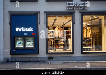 Stuttgart, 06. Januar 2022: Mode für Männer in großen Größen. Beleuchtetes Schaufenster mit Winterkleidung. Geldautomat BW Bank vor der Straße Stockfoto