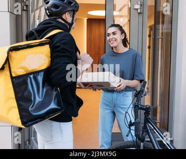 Lächelnde Frau, die ein Paket vom Kurier erhält. Lieferer mit Rucksack, der einem Kunden ein Paket in einem Wohngebäude gibt. Stockfoto