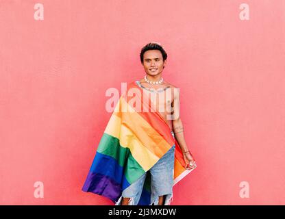 Junger, hübscher Mann, umhüllt von einer LGBT-Flagge in Regenbogenfarben an der pinken Wand Stockfoto