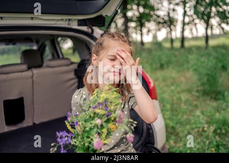 Schöne kleine Mädchen mit niedlichen Frisur sitzt im Kofferraum mit Blumenstrauß und schließen ihre Augen mit der Hand. Kind mit Sommerplan Stockfoto