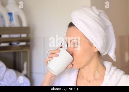 Lächelnde und frische Erwachsene Frau mit klarer Haut trägt einen Bademantel mit einem Handtuch umwickelt auf den Kopf und trinkt Kaffee im Spa zu Hause Stockfoto
