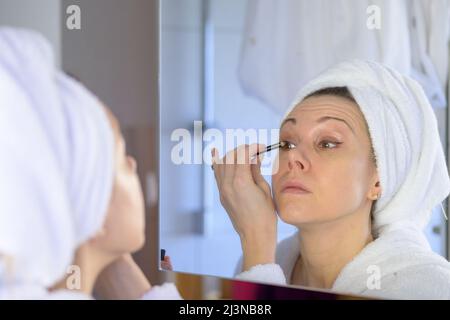 Frau mittleren Alters, die Eyeliner in ihren Badezimmerspiegel mit Fokus auf ihre Reflexion in einem Schönheits- und Pflegekonzept Stockfoto
