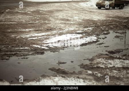 Schlammige Pfütze von geschmolzenem Schnee auf der Straße. Nicht geputzte Parkplätze. Asphalt ist mit Schneeschicht bedeckt. Stockfoto