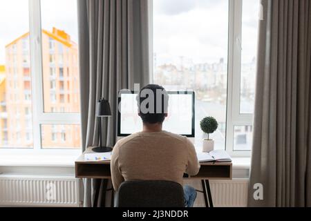 Blick von hinten auf den jungen indischen freien Mitarbeiter, der am Schreibtisch im Heimbüro sitzt, Computer benutzt, auf dem Bildschirm zuschaut, nach Geschäftsaufgaben sucht und von zu Hause aus studiert Stockfoto