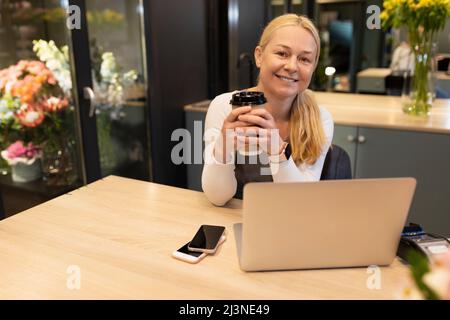 Ein Verkäufer in einem Blumengeschäft arbeitet mit einer Tasse Kaffee in den Händen an einem Computer und blickt mit einem Lächeln auf die Kamera, das Konzept eines Selbständigen Stockfoto