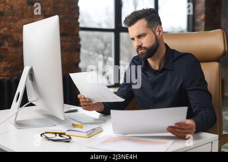 Konzentrierter kaukasischer Kerl macht Papierkram. Ein Mann schaute durch Papier und saß am Schreibtisch mit einem Computer im Büro. Man prüft das Berichtskonzept Stockfoto