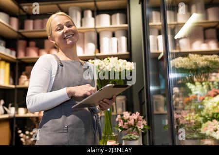 Zufriedene selbstständige Besitzerin eines Blumenladens mit einem Tablet in den Händen neben einem Kühlschrank mit frischen Blumen Stockfoto