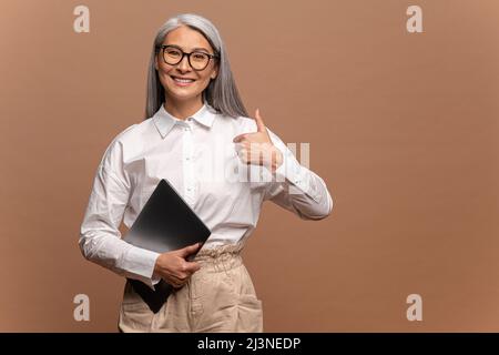 Porträt einer erfolgreichen, glücklichen Büroangestellerin, einer hübschen Frau, die steht, einen geschlossenen Laptop hält und mit toothy Lächeln auf die Kamera blickt und dabei eine coole Geste zeigt. Konzept des positiven Ausdrucks Stockfoto