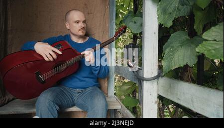 Der Mensch leitet eine Online-Akustikgitarre-Lektion von der Veranda seiner Vorstadtfarm aus Stockfoto