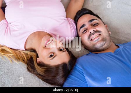 Draufsicht auf das heterosexuelle kaukasische venezolanische Paar, das im Schlafzimmer auf dem Bett liegt und die Kamera anschaut und lächelt Stockfoto