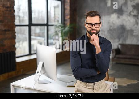 Erfolgreicher maskuliner Geschäftsmann in formell teurem Anzug, der den Arm in der Nähe des Kinn hält, hat einen ordentlichen, selbstbewussten, reichen Mann, der für Fotos posiert und die Kamera anschaut Stockfoto