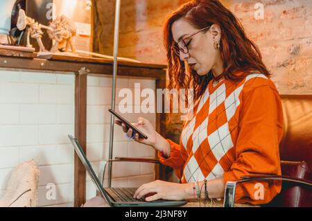 Kaukasische junge Frau, die ihren Laptop und ihr Mobiltelefon online kauft, sitzt in einem Café und macht online Geschäfte mit einer Hand auf dem Laptop ke Stockfoto