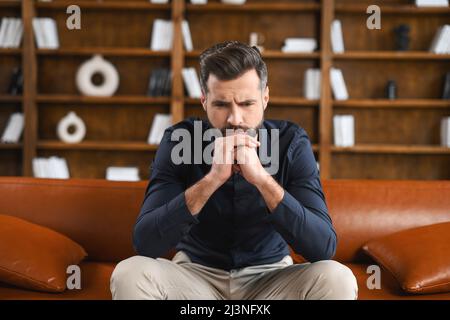 Nachdenklicher kaukasischer Kerl, der zu Hause oder im Büro auf dem Sofa sitzt. Frustrierter Mann, der sich Sorgen um die Zukunft machte, die Stirn mit der Hand bedeckte, an Probleme und Probleme nachdachte Stockfoto