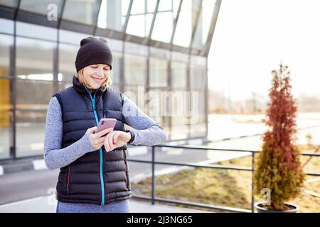 Frau mit Mobiltelefon, das an eine Smart Watch angeschlossen ist. Die Sportlerin schaut auf die Smartwatch und hält ihr Smartphone in der anderen Hand, im Freien. Fitness-FEM Stockfoto