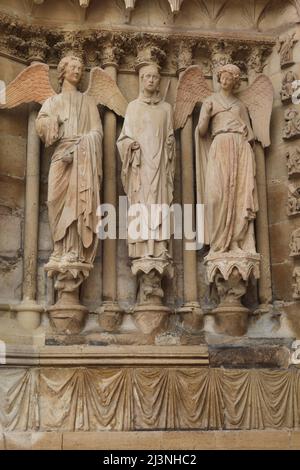 Saint Denis, umgeben von zwei Engeln, die auf dem Nordportal der Westfassade der Kathedrale von Reims (Cathédrale Notre-Dame de Reims) in Reims, Frankreich, abgebildet sind. Die berühmte gotische Statue des lächelnden Engels, die zwischen 1230 und 1245 geschnitzt wurde, ist rechts abgebildet. Die gotische Statue des Saint Denis von Paris wurde zwischen 1220 und 1230 geschnitzt, während die gotische Engelsstatue links nach 1252 geschnitzt wurde. Stockfoto