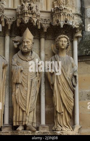 Saint Remigius von Reims auf dem zentralen Portal der Nordfassade der Kathedrale von Reims (Cathédrale Notre-Dame de Reims) in Reims, Frankreich dargestellt. Links ist der heilige Remigius abgebildet, rechts die gotische Engelsstatue. Stockfoto