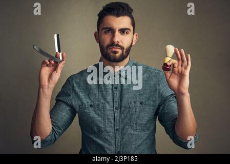 Bart wird gerade gebaut. Studioaufnahme eines hübschen jungen Mannes, der einen Rasiermesser und eine Rasierbürste in den Händen hält. Stockfoto