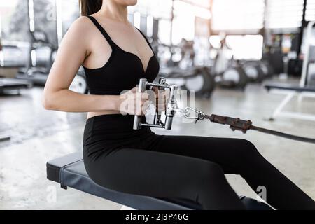 Übungskonzept die Dame trägt Sporttop, enge Hosen und schwarze Sneakers sitzen und konzentriert sich auf das Ziehen eines flexiblen Seils der Sitzrollmaschine. Stockfoto