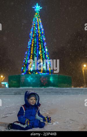 Ein kleiner Junge auf der Straße im Winter während eines Schneefalls vor einem Neujahrsbaum. Stockfoto