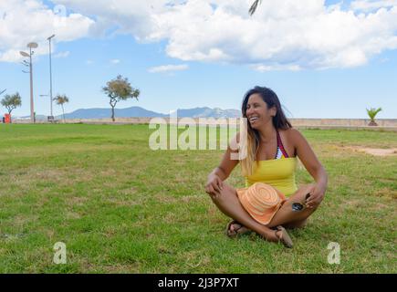 Schöne lateinische Frau 40 Jahre alt, lächelnd sitzt auf dem Rasen eines Parks auf Mallorca, balearen, hollidays Konzept Stockfoto