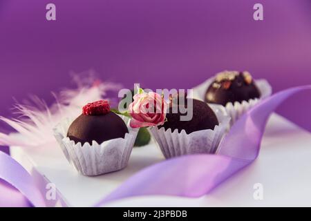 Festliche Schokoladenbonbons. Zarte rosa Federn und violettes Band. Violettes Farbmuster mit Kopierbereich Stockfoto