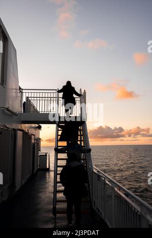 Wunderschöner Sonnenuntergang am Himmel auf der Fähre mit anonymen Passagieren, die Treppen zum oberen Deck des Schiffes hinaufsteigen. Meer und Horizont im Blick mit Licht spiegeln sich Stockfoto