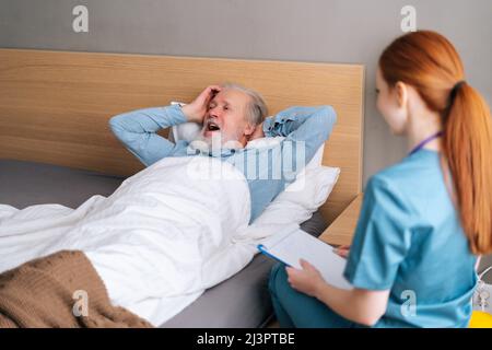 Rückansicht einer Krankenschwester, die die Diagnose und Symptome in der Krankengeschichte auf die Zwischenablage schreibt und mit einem älteren männlichen Patienten spricht, der auf dem Bett liegt Stockfoto