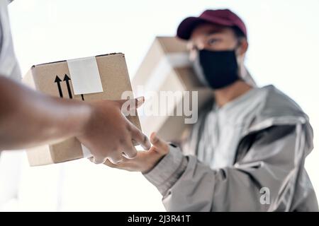 Sicher an Ihre Tür geliefert. Aufnahme eines maskierten jungen Mannes, der einem Kunden zu Hause ein Paket übergibt. Stockfoto