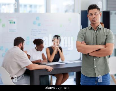 Sein Team mit größtem Engagement führen. Porträt eines selbstbewussten jungen Geschäftsmannes, der in einem Büro mit seinen Kollegen im Hintergrund steht. Stockfoto