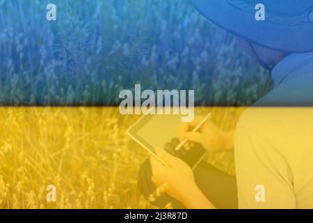 Ukraine. Digitale Farm. Porträt eines Bauern, der in einem goldenen Weizenfeld sitzt und auf einem Tablet schreibt. Junger Mann mit Cowboyhut auf dem Feld, der Weizen-cro untersucht Stockfoto