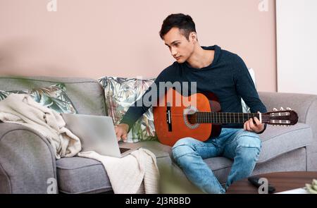 Welche Lektionen hat Lockdown gelehrt, Sie. Aufnahme eines jungen Mannes, der einen Laptop benutzt, während er zu Hause Gitarre spielt. Stockfoto