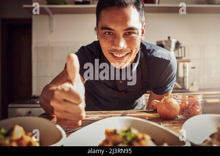 Das Essen war für einen König geeignet. Porträt eines jungen Mannes, der Daumen hoch zeigt, während er zu Hause ein köstliches Essen zubereitet. Stockfoto