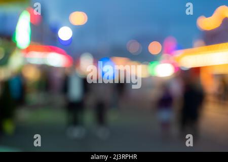 Verschwommener Hintergrund des Messegeländes mit vielen leuchtenden Lichtern. Verschwommenes Vergnügungspark mit nicht identifizierbaren Menschen wegen Bokeh. Defokussierte abstrakte Szene. Stockfoto