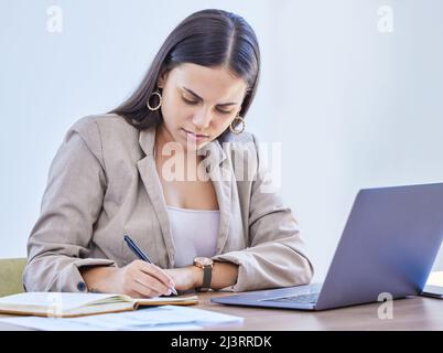 Aktualisierung ihres Tagebuchs mit einigen wichtigen Notizen. Aufnahme einer jungen Geschäftsfrau, die Notizen schreibt, während sie in einem Büro an einem Laptop arbeitet. Stockfoto
